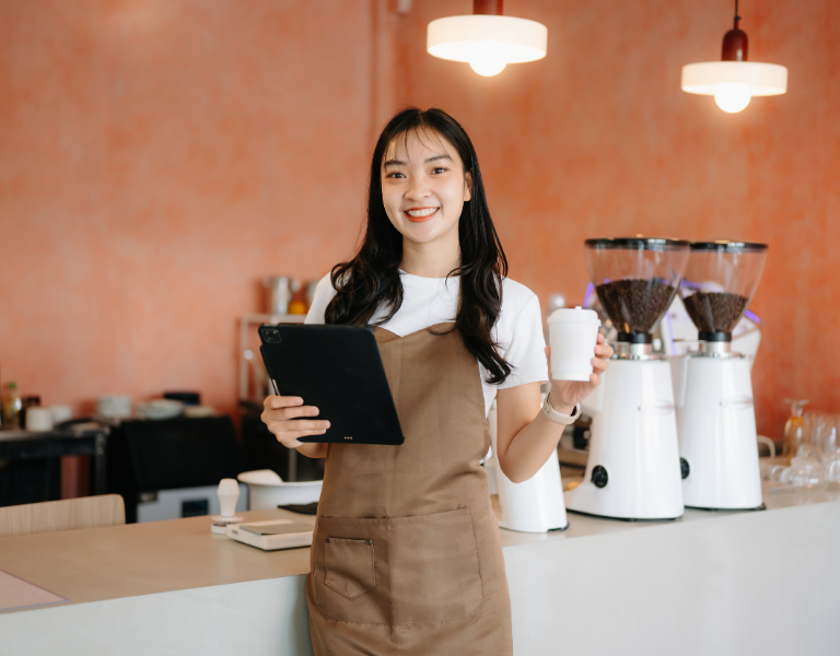 Emprendedora sonriente en su cafetería sosteniendo una tablet, simbolizando un negocio próspero con crecimiento sano y buen servicio al cliente.