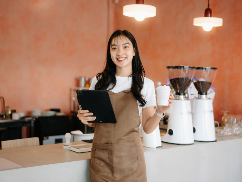 Emprendedora sonriente en su cafetería sosteniendo una tablet, simbolizando un negocio próspero con crecimiento sano y buen servicio al cliente.
